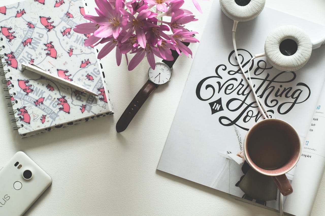 Artistic flat lay featuring a workspace with a pink-themed notebook, flowers, and headphones.
